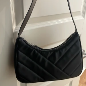 Svart quiltad axelväska - Snygg svart axelväska med quiltad design och dragkedja upptill. Väskan har en enkel och stilren look med bred axelrem, perfekt för att bära det viktigaste. Passar till många olika outfits och är lätt att matcha.