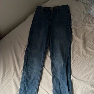 Blå jeansbyxor med normal passform - Säljer ett par klassiska blå jeansbyxor med normal passform och raka ben. Byxorna har fem fickor, knapp och dragkedja framtill samt diskreta slitningar längs benen. Perfekta till vardags och enkla att matcha med olika toppar.
