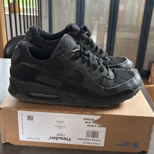 Svarta Nike Air Max 90 sneakers - Säljer ett par helsvarta Nike Air Max sneakers med klassisk design och synlig luftdämpning i sulan. Skorna har snörning, rund tå och är tillverkade i en mix av syntet och textil. Perfekta för dig som gillar streetwear och sportig stil.