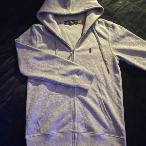 Grå hoodie från Ralph Lauren - Säljer en stilren grå hoodie från Ralph Lauren med dragkedja och broderad logga på bröstet. Tröjan har långa ärmar och en praktisk huva med snörning. Perfekt för en avslappnad look. Använt den några gånger men passar inte mig. Står att det är M men sitter som S
