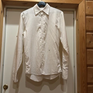 Vit skjorta från J.Lindeberg - Snygg vit skjorta från J.Lindeberg i en klassisk design. Skjortan har långa ärmar och knappar framtill. Tillverkad i en blandning av bomull och polyester för en bekväm passform. Perfekt för en stilren look.