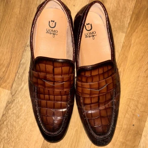 Bruna loafers från Uomo Design - Snygga bruna loafers från Uomo Design med krokodilpräglat mönster. Skorna har en elegant och klassisk design med en spetsig tå. Perfekta för en stilren look.