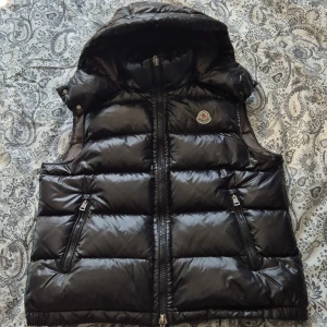 Svart dunväst från Moncler - Säljer en snygg svart dunväst från Moncler med dragkedja och huva. Västen har en glansig finish och två praktiska fickor med dragkedjor. Perfekt för kyligare dagar när du vill hålla stilen. 