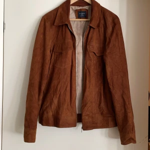 Brun mockajacka från Grand Frank - Snygg brun mockajacka från Grand Frank med dragkedja framtill. Jackan har en klassisk krage och två bröstfickor med lock. Perfekt för en stilren look.