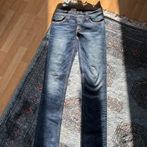 Blå jeans från Nudie Jeans Co - Säljer ett par snygga blå jeans från Nudie Jeans Co i modellen Grim Tim. De har en klassisk femficksdesign med orange sömmar och en knappgylf. Perfekta för en stilren look. (Helt nya) 