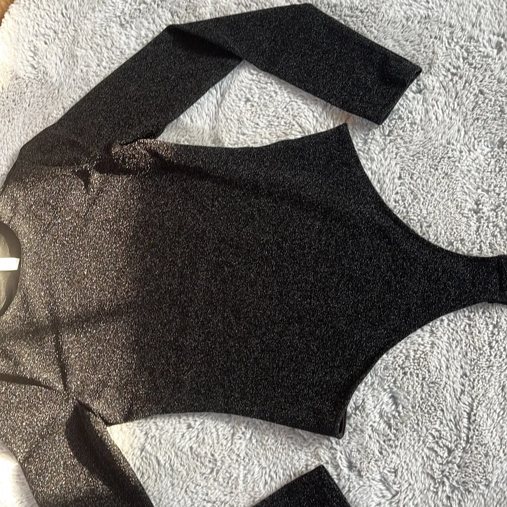 Snygg svart glittrig bodysuit med långärmad design. Perfekt för en kväll ute eller festliga tillfällen. Materialet har en skimrande effekt som fångar ljuset på ett fantastiskt sätt.. Yläosat.