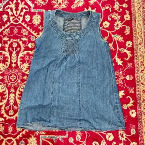 Jeanslinne  - Säljer ett snyggt jeanslinne. Linnet är ärmlöst och har en smockdetalj vid halsringningen. Perfekt för en avslappnad stil.