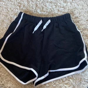 Svarta shorts med vita detaljer från H&M - Säljer ett par svarta shorts från H&M med vita kantdetaljer och snörning i midjan. Perfekta för en avslappnad stil. De har en elastisk midja för extra komfort.