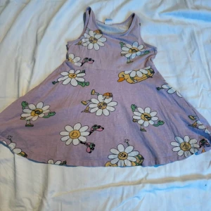 Lila klänning med blommönster - Charmig lila klänning med ett lekfullt blommönster. Klänningen är ärmlös och har en avslappnad passform, perfekt för varma dagar. Blommorna har glada ansikten och ger en rolig touch till plagget.