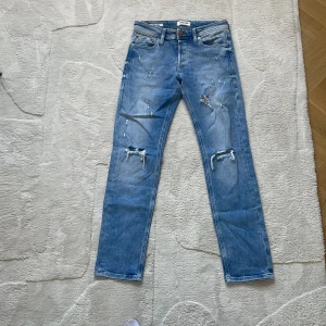 Blå jeans från Jack & Jones - Säljer ett par blå jeans från Jack & Jones med slitna detaljer och en klassisk femficksdesign. De har en knappgylf och är tillverkade i denim. Perfekta för en avslappnad stil. Modell slim Glenn  riktig schysta för sommaren!!!!