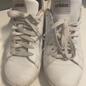 Vita sneakers från Adidas - Säljer ett par klassiska vita sneakers från Adidas med snörning. Skorna har en stilren design med perforerade detaljer på sidorna och en bekväm sula. Perfekta för en avslappnad stil.