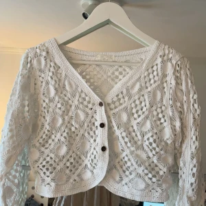 Vit spetskofta med knappar - Säljer en vacker vit spetskofta med ett unikt mönster och tre knappar framtill. Koftan har långa ärmar och en v-ringad design, perfekt för att lägga till en elegant touch till din outfit.