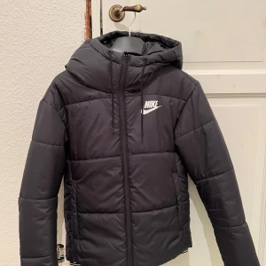 Svart dunjacka från Nike - Säljer en svart dunjacka från Nike med huva och dragkedja. Jackan har en stilren design med Nike-logga på bröstet. Perfekt för kyliga dagar. Väldigt bra skick, säljer pga för liten😀