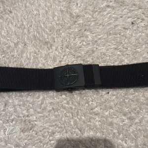 Svart bälte från Stone Island - Snyggt svart bälte från Stone Island med en stilren spänne i svart. Perfekt för att ge en cool touch till din outfit. Bältet har märkets logga både på spännet och på bandet. Skick 9/10 och 86cm långt.