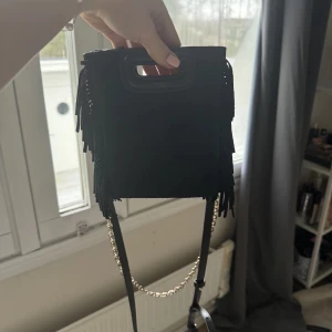 Svart axelväska med fransar - Snygg svart axelväska med fransar och kedjedetalj. Perfekt för att ge din outfit en cool touch. Väskan har ett praktiskt handtag och en justerbar axelrem.