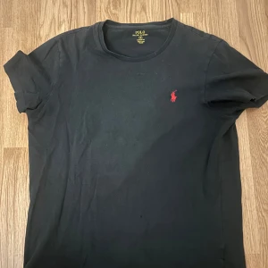Svart t-shirt från Polo Ralph Lauren - Säljer en svart t-shirt från Polo Ralph Lauren med röd broderad logga på bröstet. T-shirten har en normal passform och är kortärmad. Perfekt för en stilren och avslappnad look.
