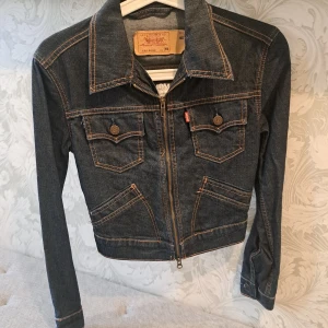 Jeansjacka från Levi's med Ramones-tryck - Säljer en cool jeansjacka från Levi's i mörk denim med ett stort vitt Ramones-tryck på ryggen. Jackan har dragkedja framtill och två bröstfickor med knappar. Perfekt för dig som älskar rock'n'roll!
