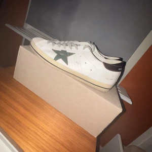 Vita sneakers med grön stjärna från Golden Goose - Snygga vita sneakers från Golden Goose med en grön stjärna på sidan. Skorna har snörning och en kontrasterande mörk häl. Perfekta för en stilren look. Kan även gå ner i pris. Skorna köptes en månad sen från merchsweden. Pris nya är 4300kr.