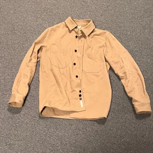 Beige overshirt med knappar - Snygg beige overshirt med långa ärmar och knappar framtill. Skjortan har två bröstfickor och en klassisk krage. Perfekt för lager-på-lager-stil.  Kommer ej ihåg vart den är köpt eller hur mycket den är köpt för och har aldrig använt den, den har bara legat gömd i min garderob och passar inte mig längre.