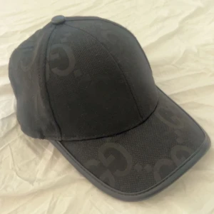 Svart gucci keps - Snygg svart keps från Gucci med diskret mönster av märkets logotyp. Tillverkad i Italien med högkvalitativt bomullstyg. Perfekt för en stilren look. Inget tillbehör kommer med , drf säljer jag den vid ett hyfsat bra pris , kepsen är helt ny med