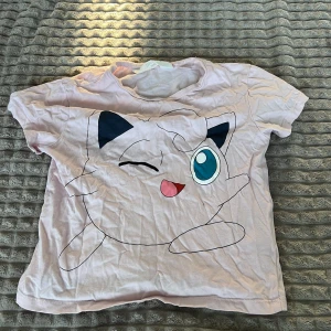 Rosa Pokémon t-shirt - Säljer en söt rosa Pokémon t-shirt med en stor Jigglypuff-tryck på framsidan. T-shirten är kortärmad och perfekt för alla Pokémon-fans. Den har en avslappnad passform och är gjord i mjukt material för extra komfort.