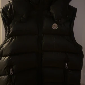 Svart dunväst från Moncler - Säljer en stilren svart dunväst från Moncler med dragkedja och hög krage. Västen har en quiltad design och är perfekt för kyligare dagar. Den har även Monclers logotyp på bröstet.