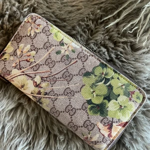 Blommig plånbok med dragkedja - Snygg plånbok med blommigt mönster i gröna och beige toner. Plånboken har en dragkedja i guld och flera fack för kort och kontanter. Perfekt för att hålla ordning på dina tillhörigheter med stil.