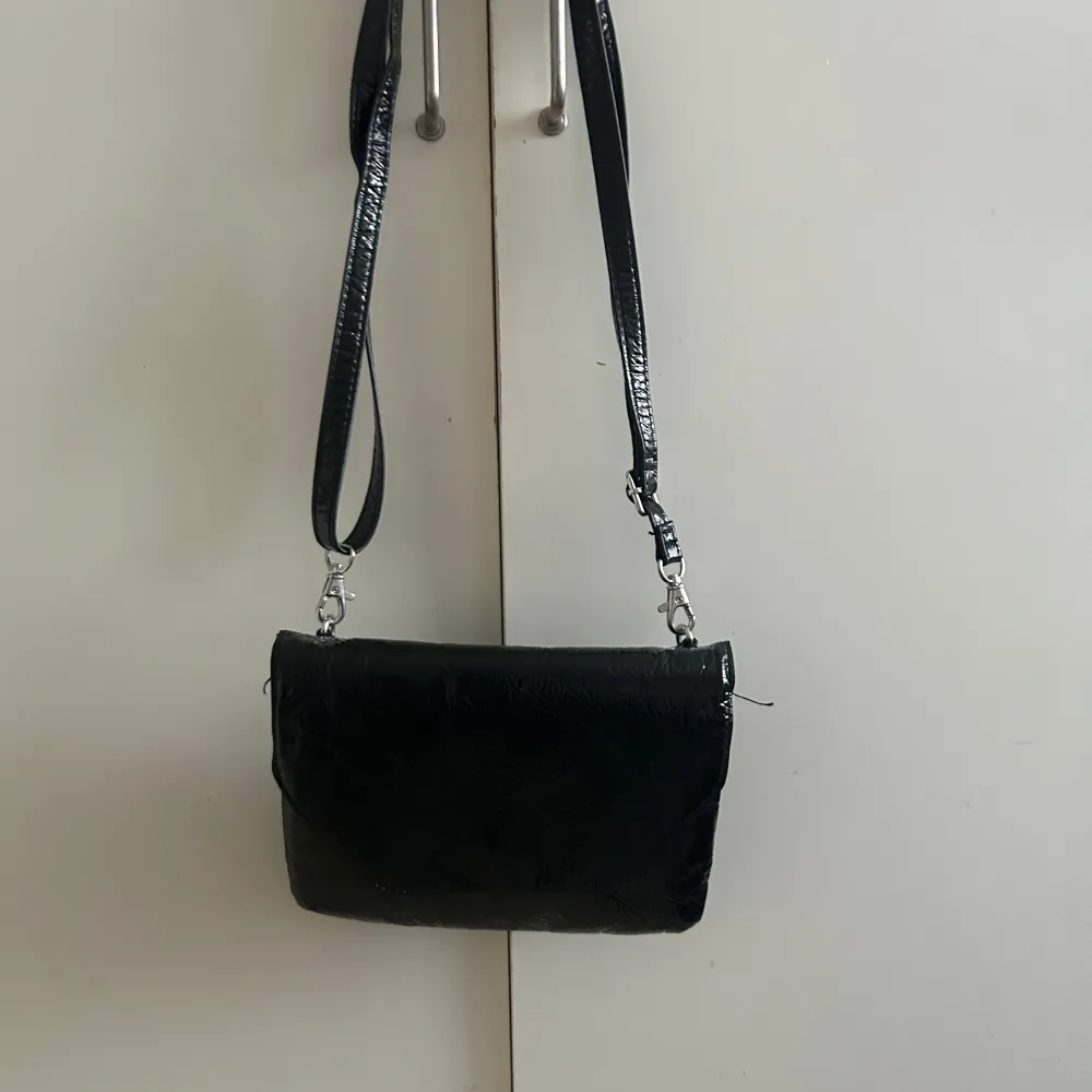 Snygg svart axelväska från Monki med justerbar rem. Väskan har en stilren design och är perfekt för att bära dina nödvändigheter. Den är tillverkad i ett glansigt material som ger en modern touch.. Laukut & Käsilaukut.