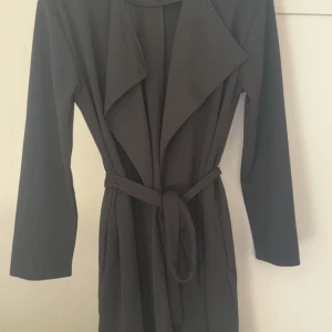 Grå trenchcoat från Vila - Snygg svart  trenchcoat från Vila med knytbälte i midjan. Jackan har långa ärmar och en klassisk krage. Perfekt för en stilren look.