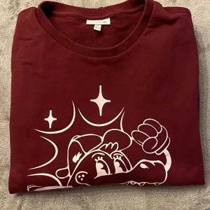 Vinröd tröja från Lacoste med tryck - Säljer en vinröd tröja från Lacoste med ett stort tryck av en tecknad figur och Lacoste-loggan på framsidan. Tröjan har långa ärmar och ribbade muddar vid ärmslut och nederkant. Perfekt för en avslappnad stil!