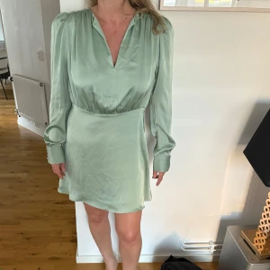 Mintgrön kort klänning i satin - Superfin mintgrön klänning i satin med lång ärm och v-ringning. Klänningen har markerad midja och lätt puff vid axlarna för en snygg siluett. Perfekt för dig som gillar en stilren look med lite extra shine.