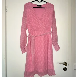 Rosa omlottklänning SisterS Point - Supersöt rosa kortklänning från SisterS Point med vita prickar, omlottmodell och knytband i midjan. Långa ärmar med manschett och v-ringad hals. Perfekt för dig som gillar en feminin och lekfull stil.