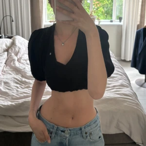 Svart blus med knyt i ryggen - Supersöt svart croppad blus med puffiga korta ärmar och v-ringning framtill. Toppen har öppen rygg med knytband och är gjord i ett mjukt, strukturerat bomullstyg. 