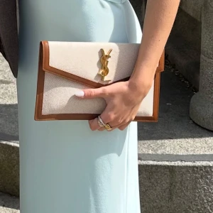 Saint Laurent clutch  - Säljer min snygga clutch från Saint Laurent i beige canvas med bruna skinnkanter och guldigt YSL-emblem framtill. Använd ca 3 gånger och är i super skick. Köpt förra året. Kvitto och äkthetsbevis samt låda/box medföljer. Mini fläck där bak längst ned men inget som syns (sista bilden). 