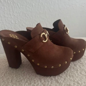 Nya. Bruna mocka träklackar från San Marina - Chunky träklackar i brun skinn/mocka från San Marina med guldfärgade metalldetaljer och nitar längs sidorna. Skorna har hög klack och plattform, rund tå och slip-in modell. Perfekta för dig som vill sticka ut med retrovibbar och coola detaljer.