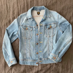Ljusblå jeansjacka från Lee - Klassisk ljusblå jeansjacka från Lee med snygga kontrastsömmar och bruna knappar. Jackan har två bröstfickor med lock och knapp, samt justerbara knappar i midjan. Perfekt för dig som gillar en tidlös och avslappnad stil.