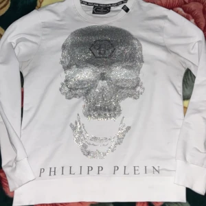 Vit Philipp Plein tröja med glitterdödskalle - Säljer en vit långärmad tröja från Philipp Plein med en stor glittrig dödskalle i silver på bröstet och logotypen tryckt nedtill. Tröjan har rund hals och är tillverkad i mjuk bomull. Perfekt för dig som gillar statement-plagg och exklusiv streetstyle.