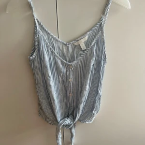 Randig linnetopp med knyt från H&M - Superfin blåvit randig linnetopp från H&M med smala axelband, knappar framtill och knyt längst ner. Perfekt för varma dagar och ger en chill sommarkänsla. Tunt och luftigt material som känns skönt mot huden