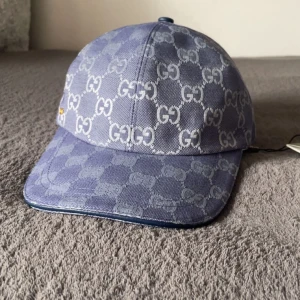 Gucci keps Dubai blå - Snygg blå keps från Gucci med ikoniskt GG-mönster över hela ytan. Justerbar rem baktill i blått och detaljer i guld. Kepsen är tillverkad i bomull och har en klassisk böjd skärm. Perfekt för dig som vill sticka ut med en lyxig accessoar.