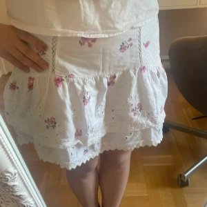 Vit blommig volangkjol med spets - Supersöt vit kortkjol med rosa blommor, volanger och broderade spetsdetaljer. Kjolen har en romantisk vibe med brodyr och små hålmönster, samt dekorativa sömmar och spetsband. Perfekt för dig som gillar en feminin och somrig stil.