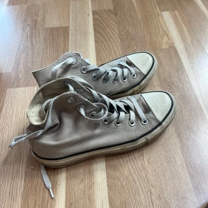 Beige Converse - Säljer ett par klassiska Converse All Star high tops i beige canvas med vita snören och rund tå. Skorna har den ikoniska loggan på sidan och en platt sula med svart rand. Perfekta för en avslappnad och cool streetstil.