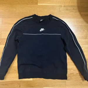 Svart Nike sweatshirt med vita detaljer - Snygg svart sweatshirt från Nike med vita ränder längs ärmarna och en vit logga på bröstet. Tröjan har rund halsringning och långa ärmar. Perfekt för en sportig och avslappnad stil.