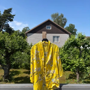 Gul blommig skjorta från Pull&Bear - Snygg gul skjorta från Pull&Bear med blommigt mönster och knytband i midjan. Skjortan har lös passform och trekvartsärmar, perfekt för soliga dagar. Materialet känns svalt och lätt, vilket gör den superbekväm att bära. Passar xs-m