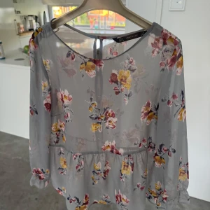 Blommig blus från Zara Trafaluc S - Supersöt blommig blus från Zara Trafaluc i ljusgrått med gula, rosa och vinröda blommor. Blusen är tunn och luftig med långa ärmar, volang nertill och knappar i ryggen. Perfekt för dig som gillar romantisk och feminin stil.