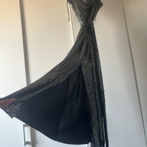 Glittrig långklänning med fransar - Säljer en svart glittrig långklänning med fransdetaljer i midjan från ZARA. Klänningen har en snygg omlott-look och är ärmlös med en elegant siluett. Perfekt för dig som vill sticka ut med en glamorös vibe på nästa fest eller bal.