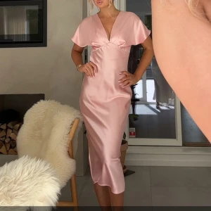 Ljusrosa satinklänning med v-ringning - Superfin ljusrosa långklänning i satin med v-ringad hals och korta ärmar. Klänningen har en elegant, figurnära passform och ett mjukt, glansigt material som ger en lyxig känsla. Perfekt för dig som vill sticka ut med en romantisk och trendig look.