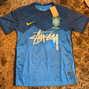 Stüssy Nike fotbollströja - Snygg blå Brasil fotbollströja från Nike i samarbete med Stüssy. Tröjan har korta ärmar, Brasil-logga på bröstet, gul Nike swoosh och Stüssy-tryck framtill. Tillverkad i Dri-FIT-material som andas, perfekt för träning eller match.