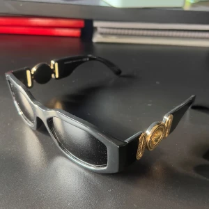 Svarta Versace solglasögon med guld ton - Snygga svarta solglasögon från Versace med rektangulär form och ikoniska gulddetaljer med Medusa-logga på skalmarna. Glasögonen har en chunky design och levereras med originalfodral och certifikat. Perfekt accessoar för dig som vill sticka ut.