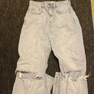 Ljusblå slitna jeans med hål - Säljer ett par ljusblå jeans med rejält slitna knän och fransiga detaljer. Klassisk femficksmodell med hög midja och raka ben. Perfekta för en avslappnad och trendig streetstil.