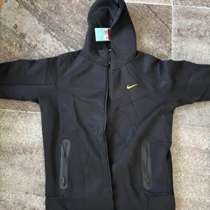 Svart Nike NOCTA hoodie med dragkedja - Svart hoodie från Nike NOCTA med gul Swoosh på bröstet och NOCTA-tryck på ärmen. Jackan har hel dragkedja, två fickor med dragkedja och en skön huva. Tillverkad i mjuk bomullsmix, perfekt för chill eller träning. Storlek L men passar som M